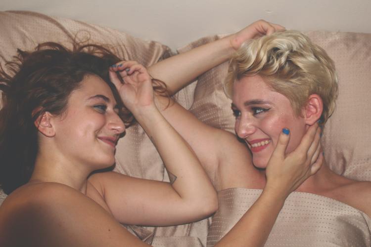 Two smiling women are in bed together. One woman touches the other woman's cheek.